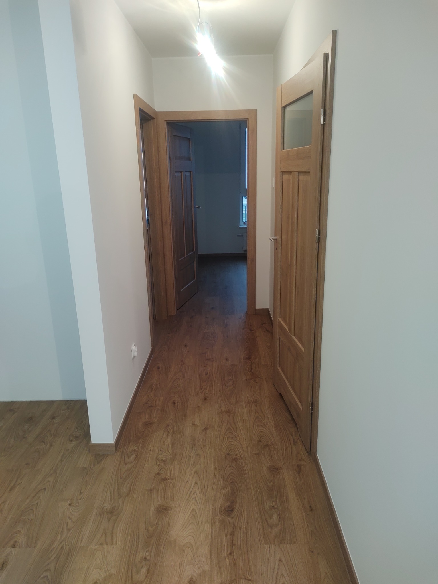 Korytarz po remoncie z panelami podłogowymi w odcieniach brązu i białymi ścianami. Widoczne dwoje drewnianych drzwi, jedne z przeszkleniem. Oświetlenie sufitowe.