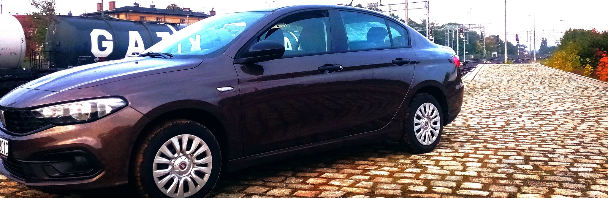 Ciemnobrązowy samochód osobowy marki Fiat stojący na brukowanej nawierzchni peronu kolejowego, w tle wagony cysterny z napisem GAZ i sygnalizacja świetlna.