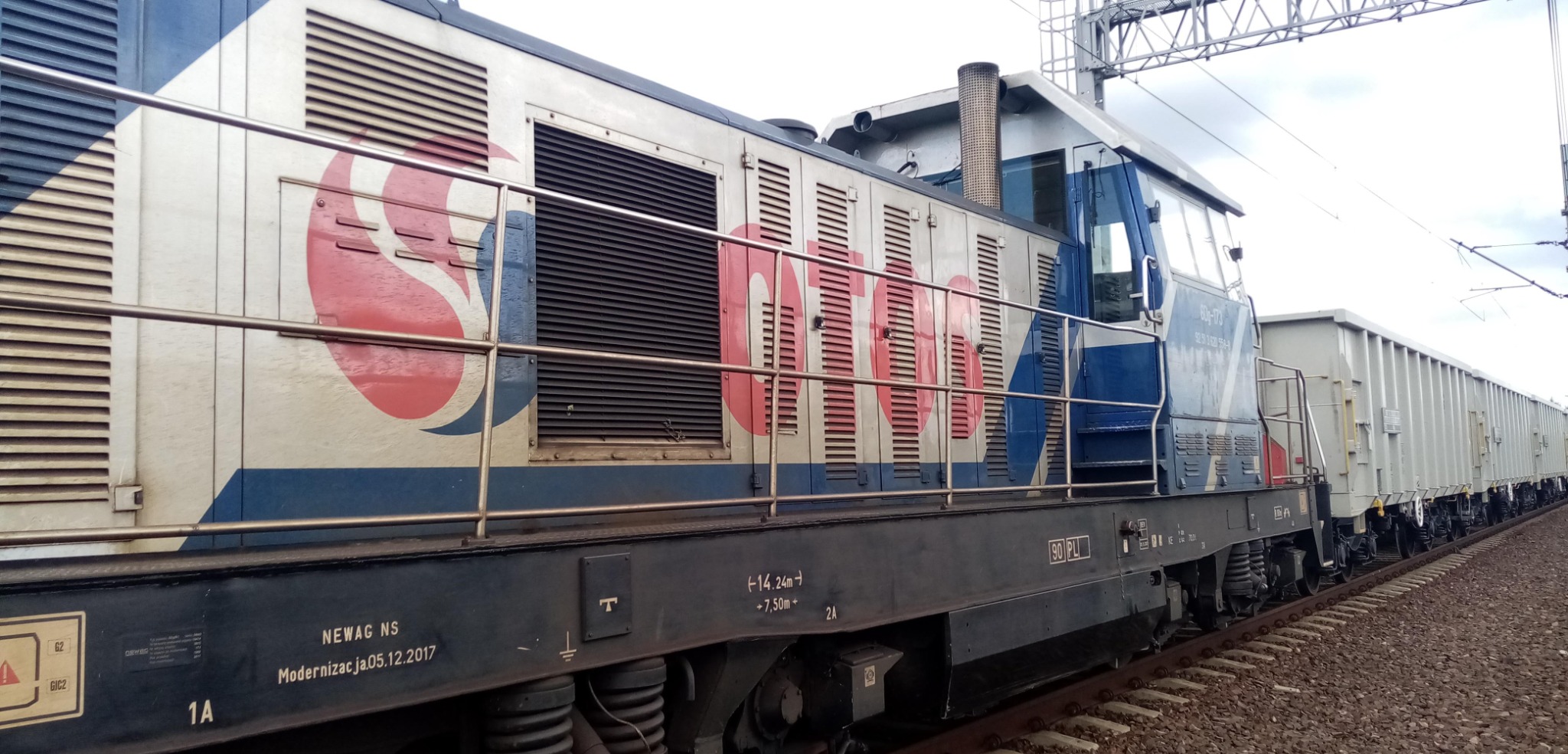 Bok lokomotywy NEWAG NS z wagonami towarowymi na torach kolejowych, widoczne detale malowania i oznaczenia techniczne.