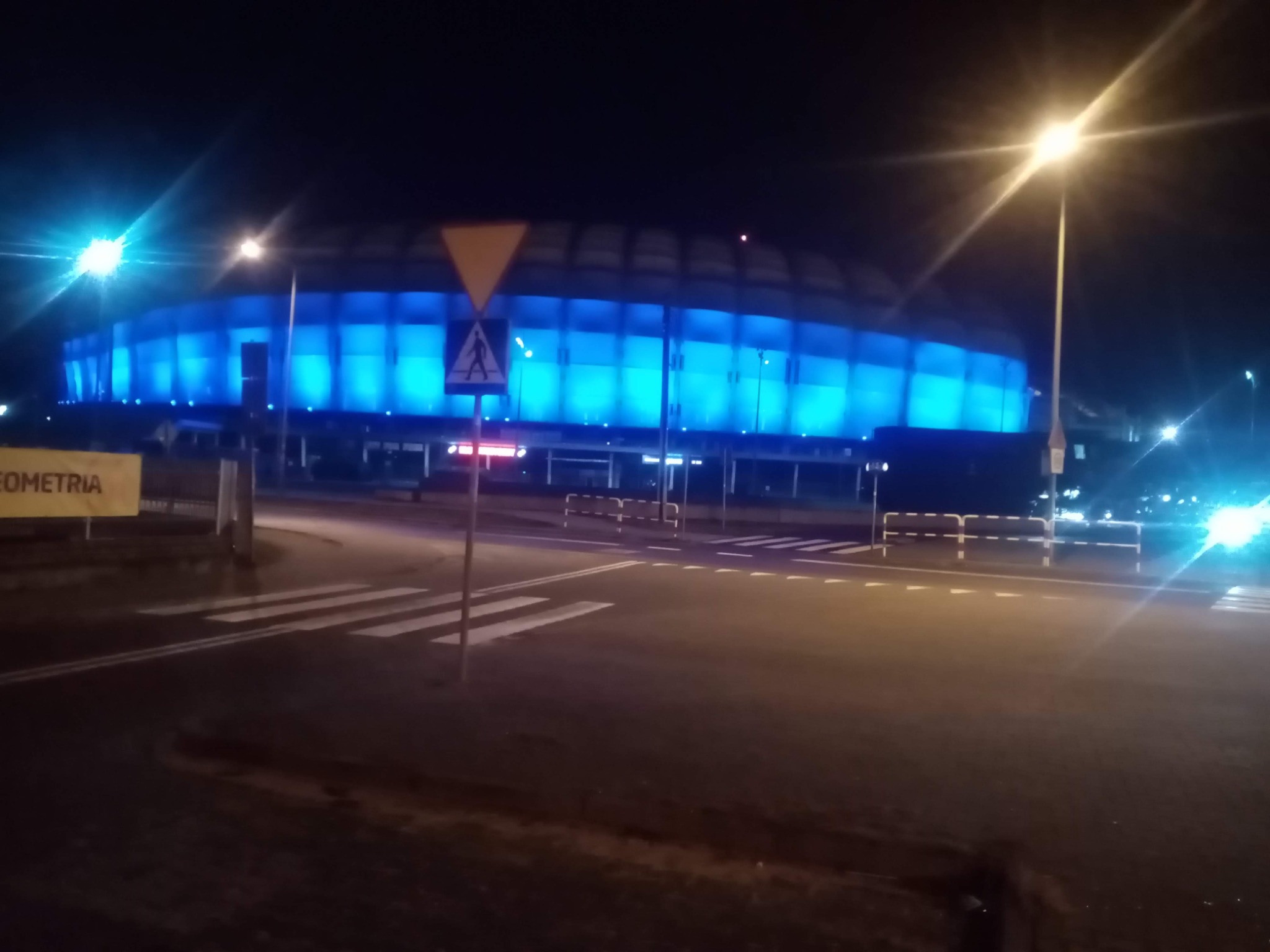 Nocne ujęcie stadionu oświetlonego na niebiesko, widok z ulicy z przejściem dla pieszych i znakiem drogowym.