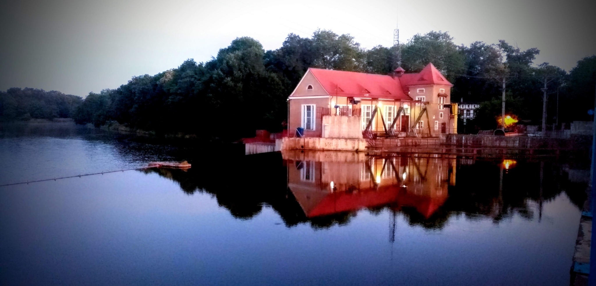 Mała elektrownia wodna z czerwoną dachówką, odbijająca się w spokojnej tafli rzeki, otoczona zielenią drzew.