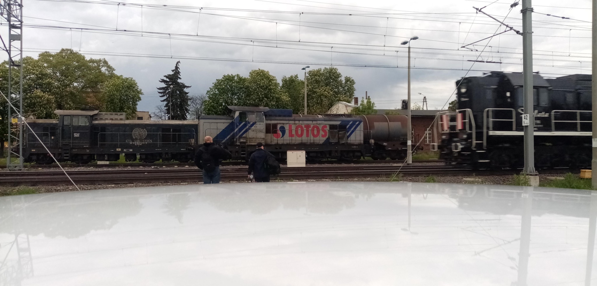 Dwa lokomotywy kolejowe, jeden z logo LOTOS, stojące na torach kolejowych z dwoma osobami w pobliżu, widoczne zza białej, błyszczącej powierzchni.