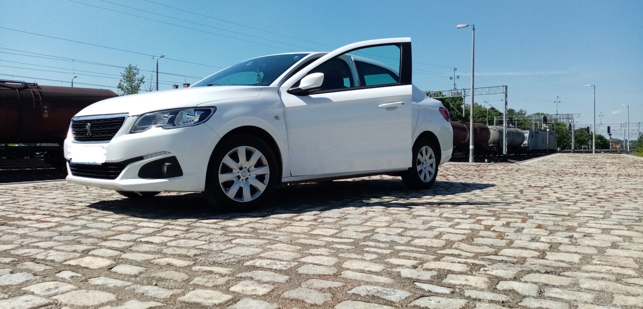 Biały samochód osobowy z otwartymi drzwiami pasażera, zaparkowany na brukowanej nawierzchni w pobliżu torów kolejowych i wagonów cystern.