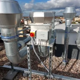 Metalowe rury wentylacyjne i skrzynka sterownicza z czerwonym przyciskiem na dachu budynku z widokiem na miasto w tle.