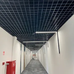Długi korytarz biurowy z podwieszanym, czarnym sufitem rastrowym i nowoczesnym oświetleniem LED wzdłuż ścian. Na ścianie po lewej stronie widoczna czerwona szafka hydrantowa.