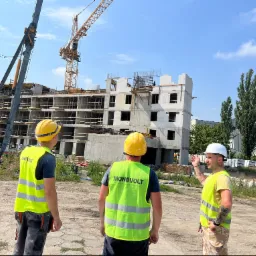 Trzech pracowników w kamizelkach odblaskowych i kaskach na placu budowy w trakcie wykonywania instalacji elektrycznych w budynku wielorodzinnym, w tle widoczny żuraw budowlany.