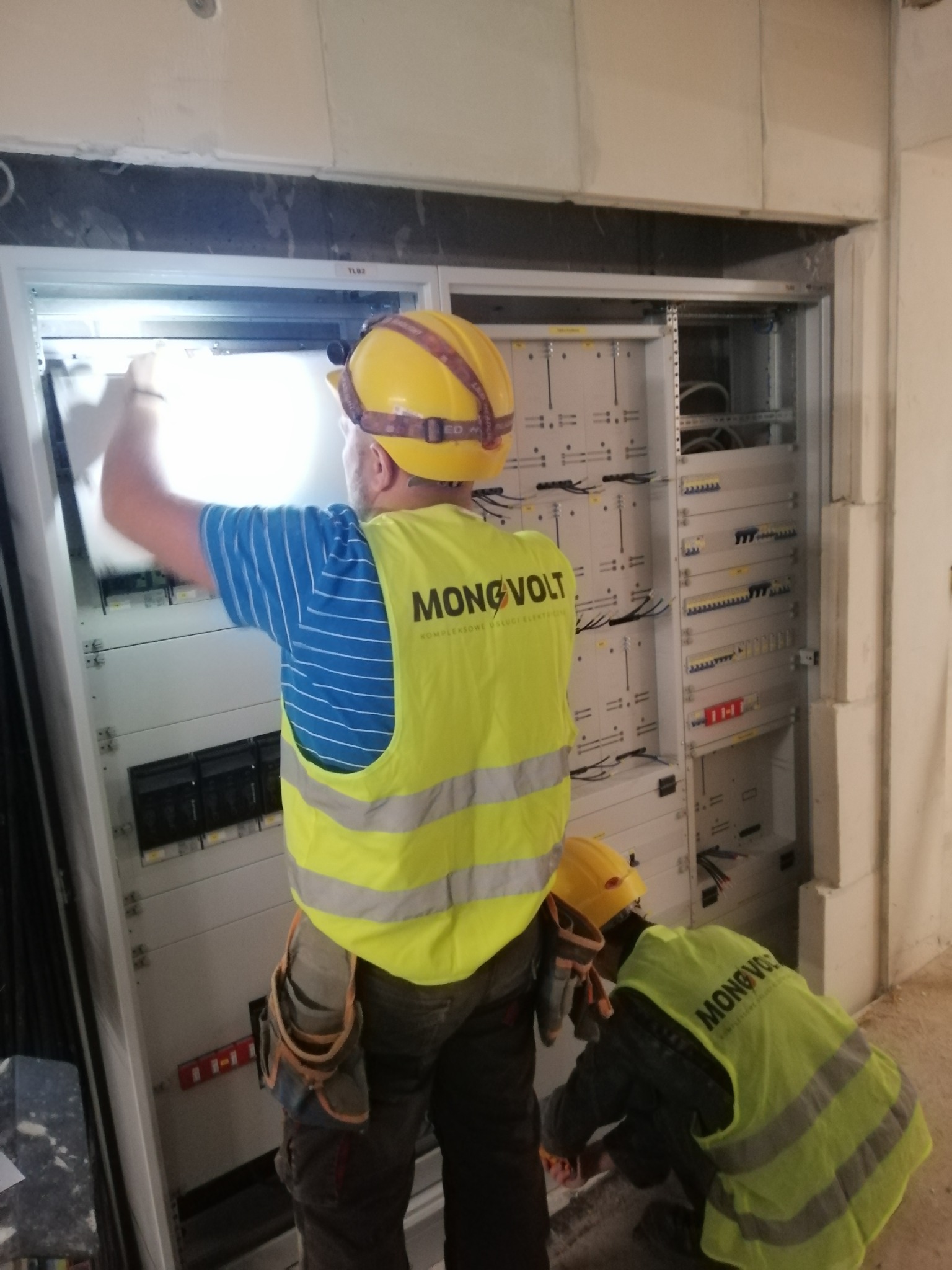 Dwóch elektryków w żółtych kamizelkach z logo firmy pracuje przy montażu rozdzielnicy elektrycznej w surowym wnętrzu, jeden z nich oświetla sobie wnętrze szafy rozdzielczej latarką czołową.
