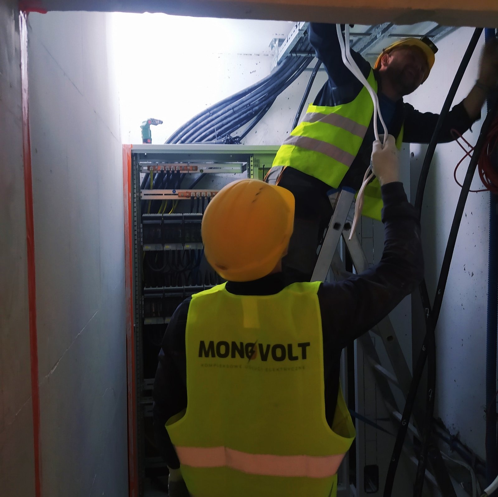 Dwóch elektryków w żółtych kamizelkach i kaskach, jeden na drabinie, drugi stojący, prowadzą przewody w pobliżu rozdzielnicy elektrycznej w wąskim pomieszczeniu technicznym.