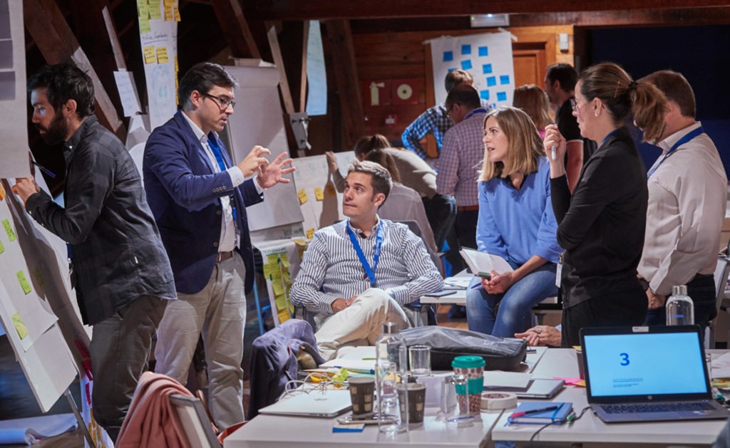 Grupa osób w trakcie burzy mózgów w przestrzeni biurowej na poddaszu, mężczyzna w niebieskiej marynarce gestykuluje podczas prezentacji, inni uczestnicy słuchają i notują, laptop z numerem 3...