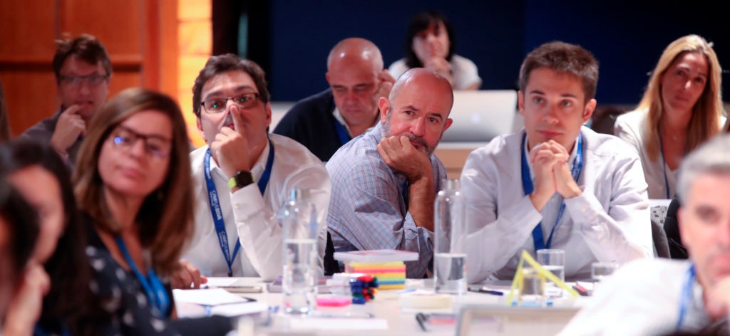 Grupa osób uczestniczy w seminarium lub szkoleniu biznesowym, siedząc przy stole z notatkami, długopisami i butelkami wody.