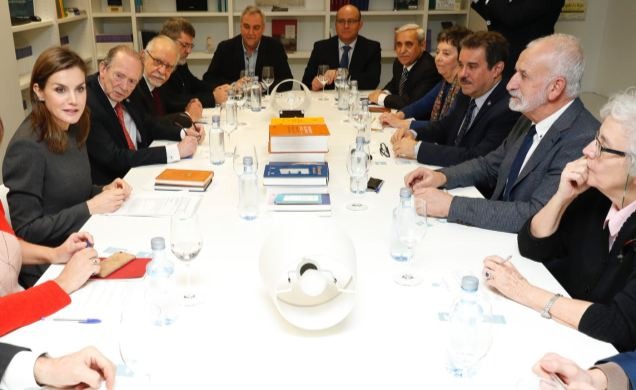 Grupa osób w garniturach siedzi przy stole konferencyjnym z notatnikami, książkami i butelkami wody, w tle regał z książkami.