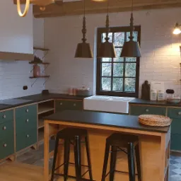 Rustykalna kuchnia z zielonymi szafkami, ciemnym blatem, białymi płytkami na ścianach i drewnianą wyspą z czarnymi stołkami barowymi.