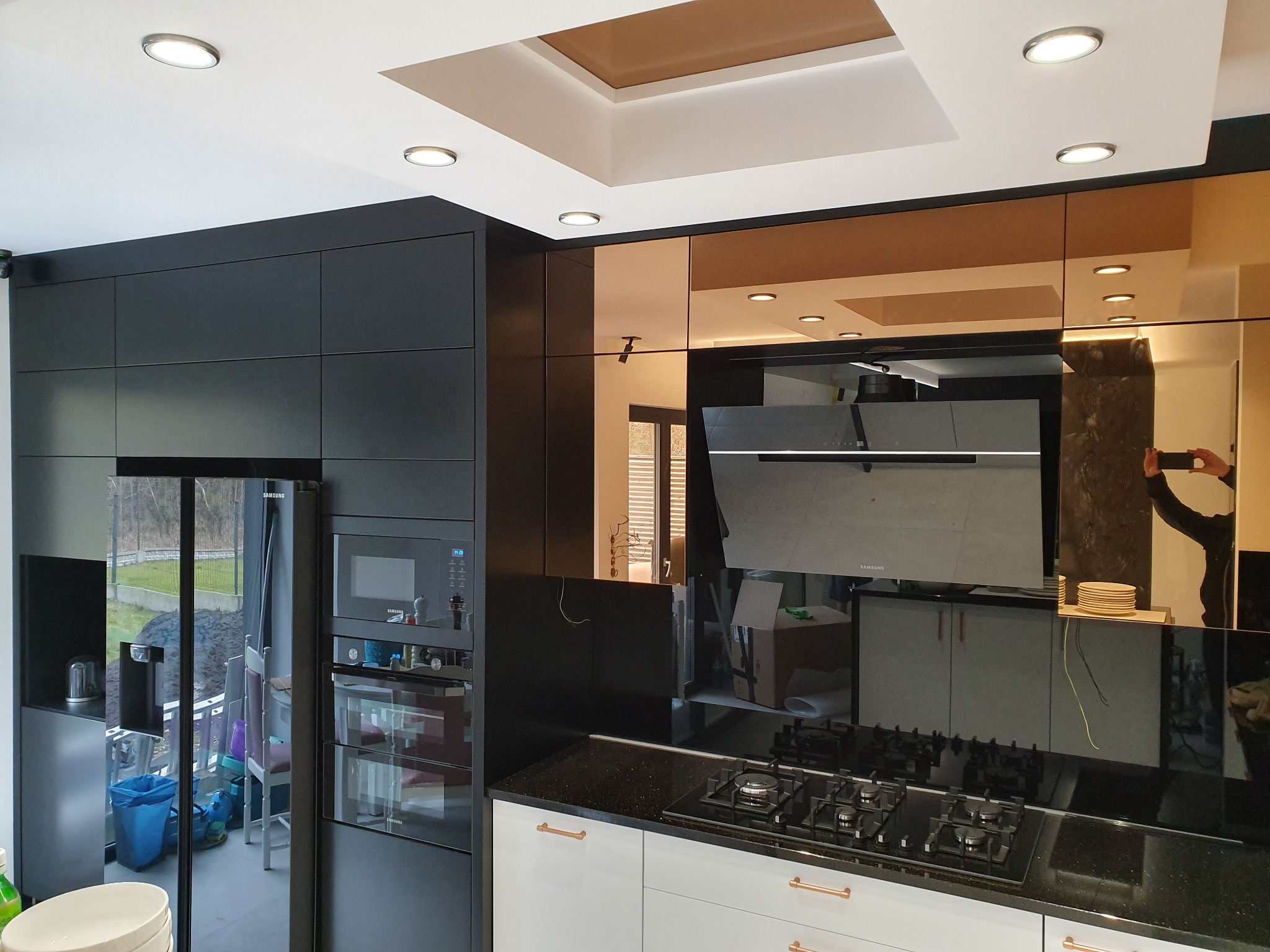 Nowoczesna kuchnia z czarnymi i białymi szafkami, blatem z czarnego granitu, złotymi elementami dekoracyjnymi i lustrzanymi panelami ściennymi. Widoczny fragment osoby robiącej zdjęcie w odbiciu.