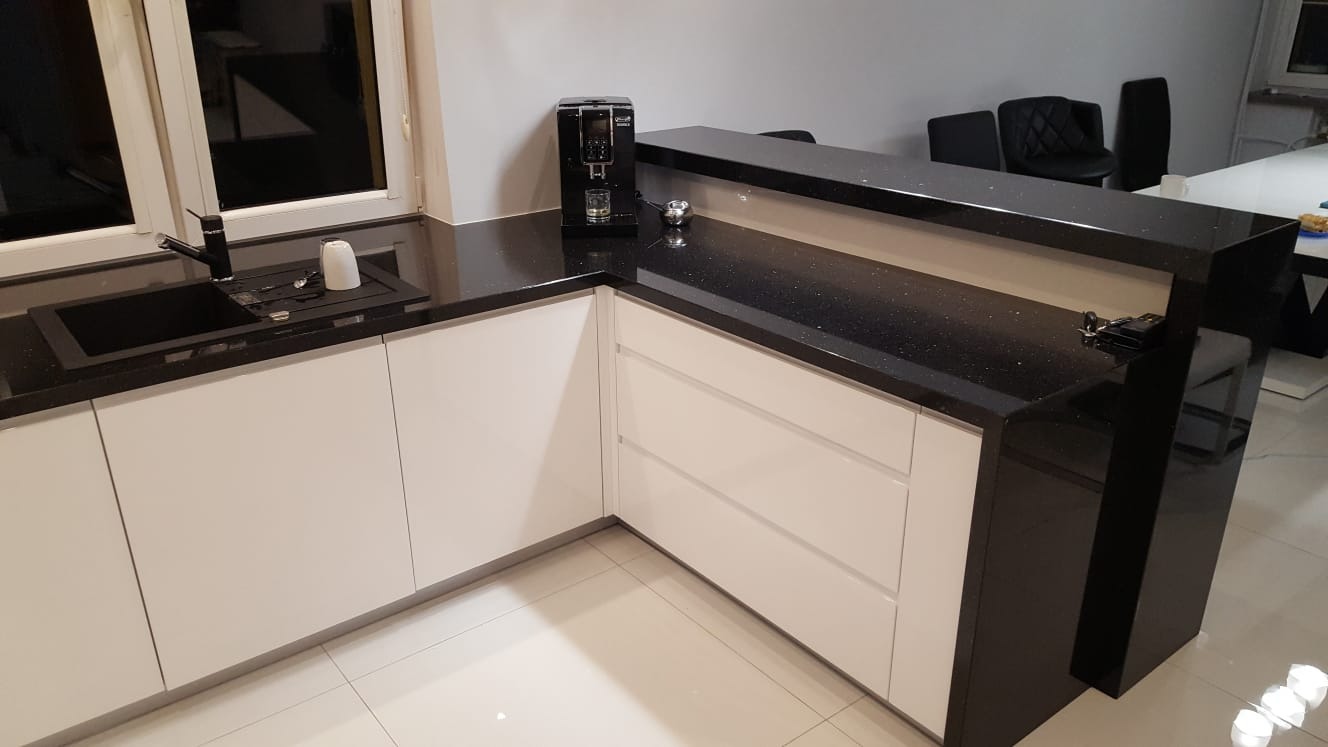 Nowoczesna kuchnia z białymi frontami i czarnym blatem z efektem gwiazd, wyposażona w czarny zlew i ekspres do kawy.