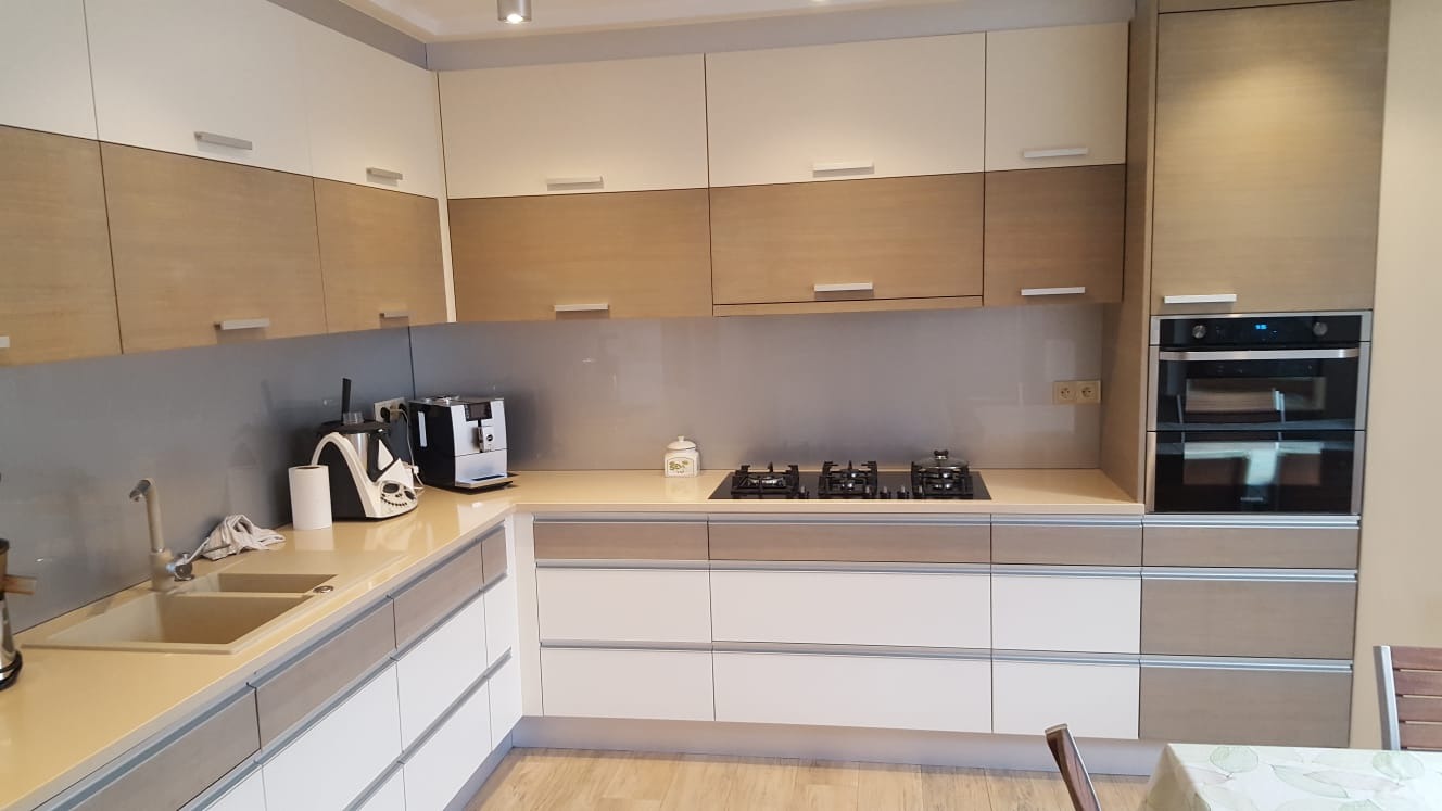 Nowoczesna kuchnia z dwukolorowymi szafkami (białe i drewnopodobne), szarym szkłem lacobel nad blatem, płytą indukcyjną i piekarnikiem w zabudowie.