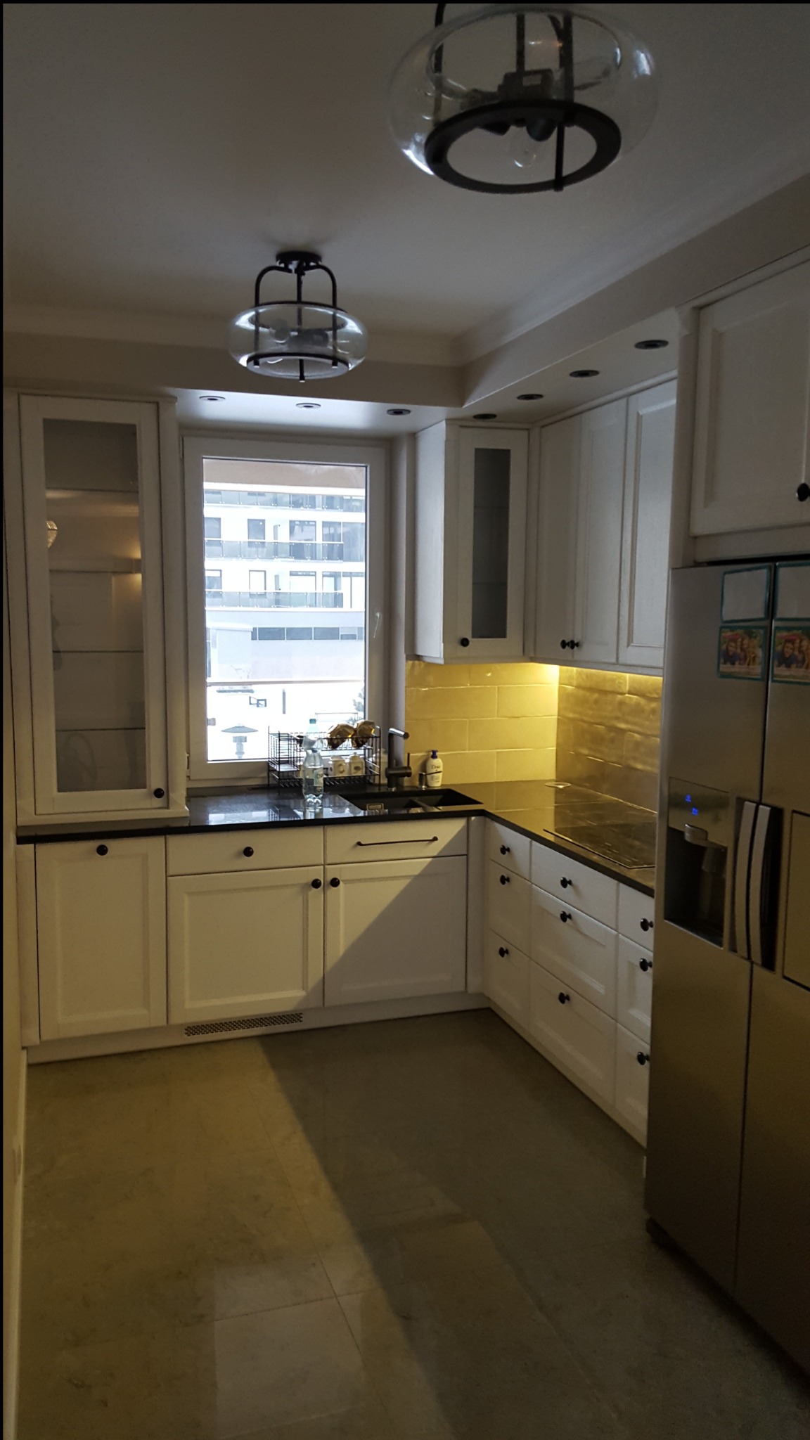 Jasna kuchnia z białymi szafkami, czarnymi blatami i złotą mozaiką na ścianie, oświetlona lampami sufitowymi w stylu industrialnym.