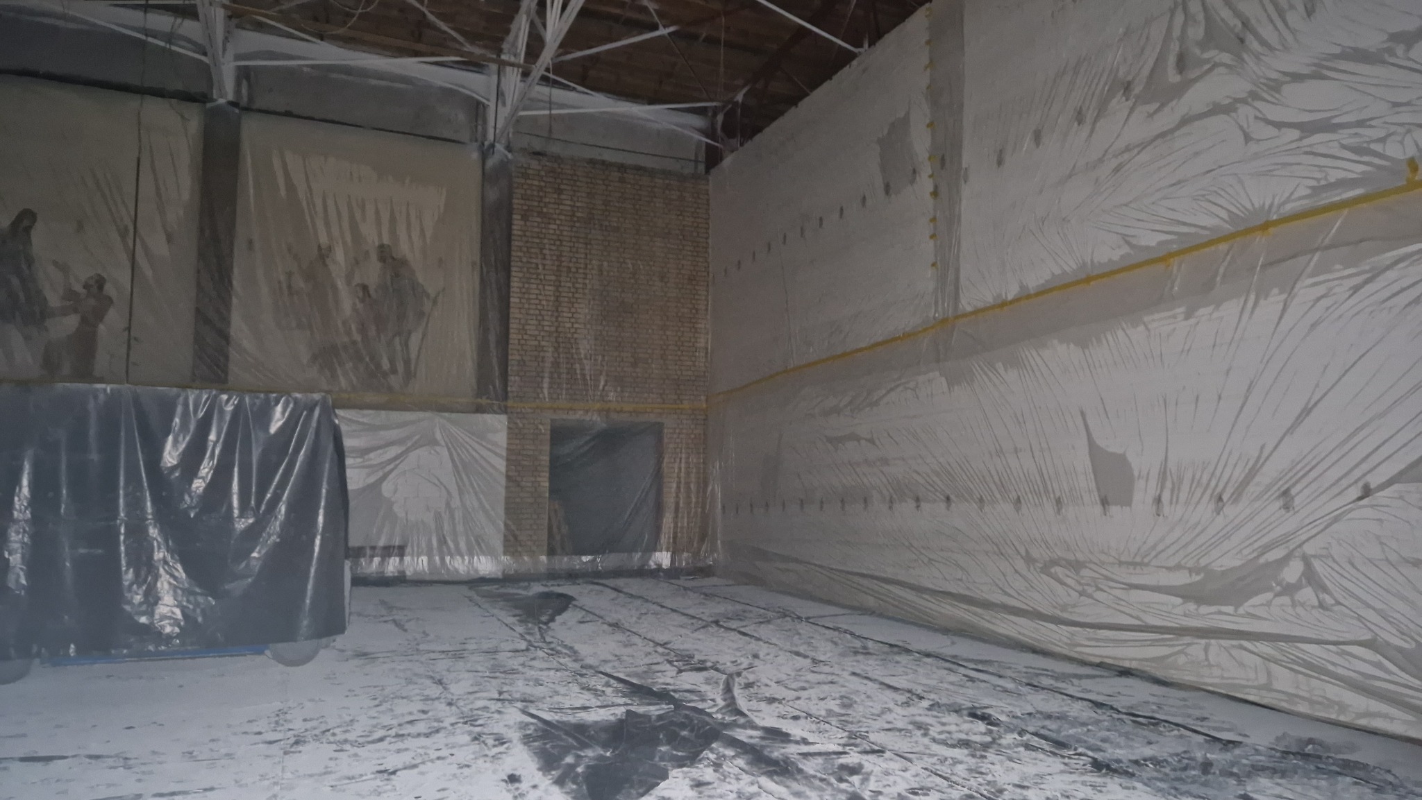 Duża hala przygotowana do prac remontowych, zabezpieczona folią malarską, widoczne zasłonięte malowidła na ścianach.