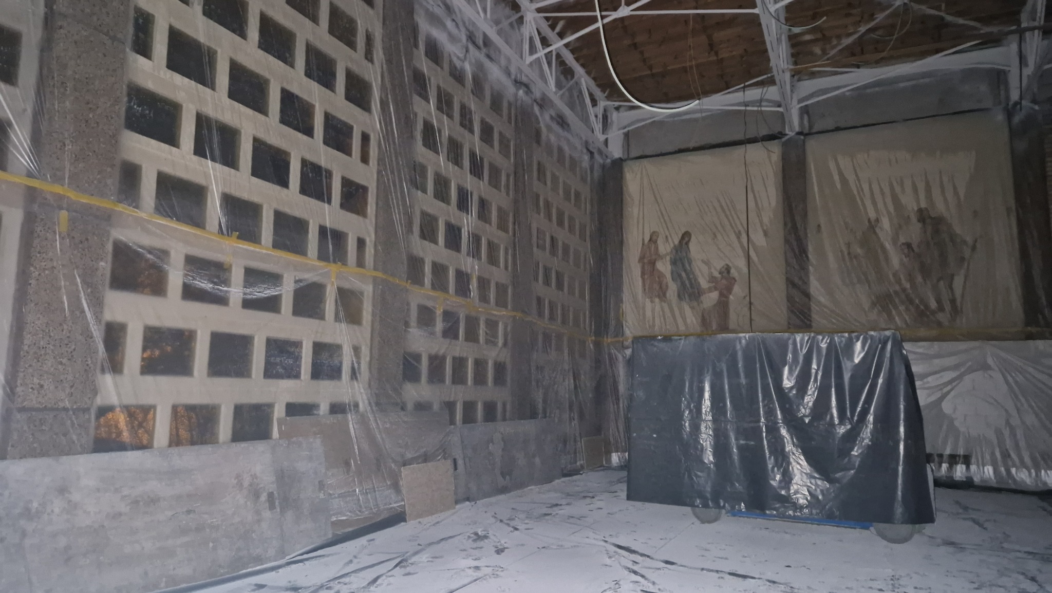 Wnętrze pomieszczenia zabezpieczonego folią malarską przed pracami remontowymi; widoczne ściany z charakterystycznymi otworami oraz malowidła ścienne również zabezpieczone folią; podłoga pokryta...