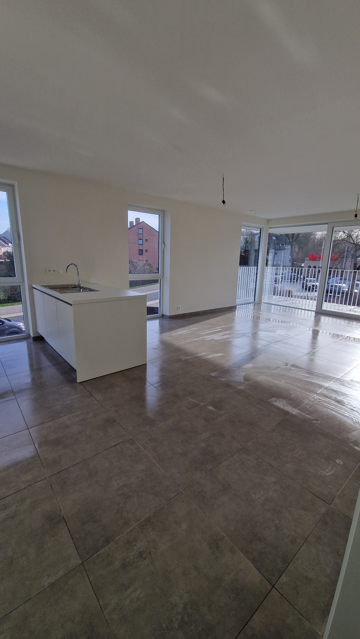 Wnętrze jasnego, przestronnego mieszkania z dużymi oknami i szarą, błyszczącą podłogą, widoczna biała wyspa kuchenna z kranem, ślady po myciu podłogi.