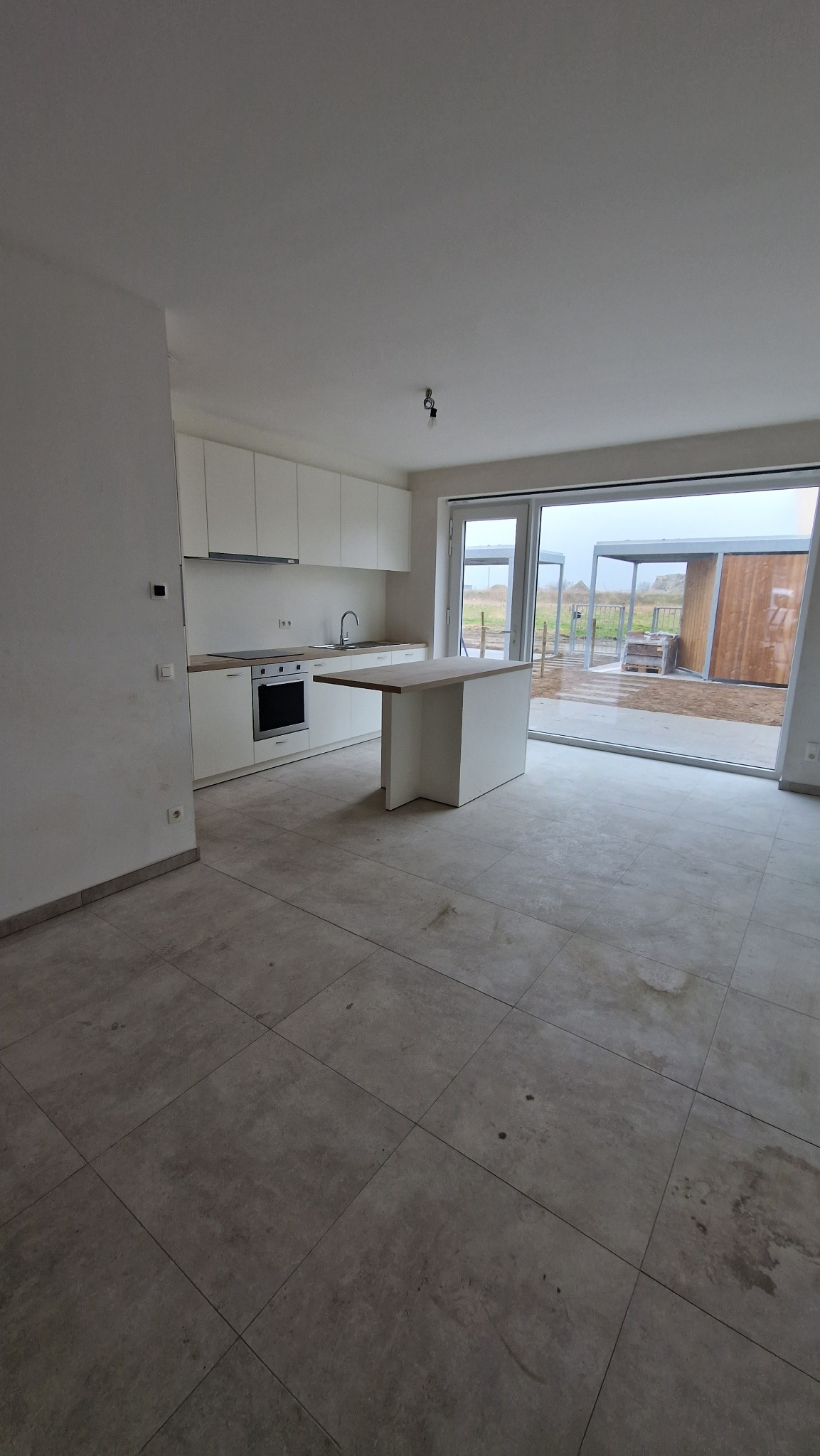 Wnętrze surowego mieszkania z widoczną białą zabudową kuchenną, wyspą z drewnianym blatem i szarą, nie do końca czystą podłogą z płytek, duże przesuwne okno z widokiem na zewnątrz.