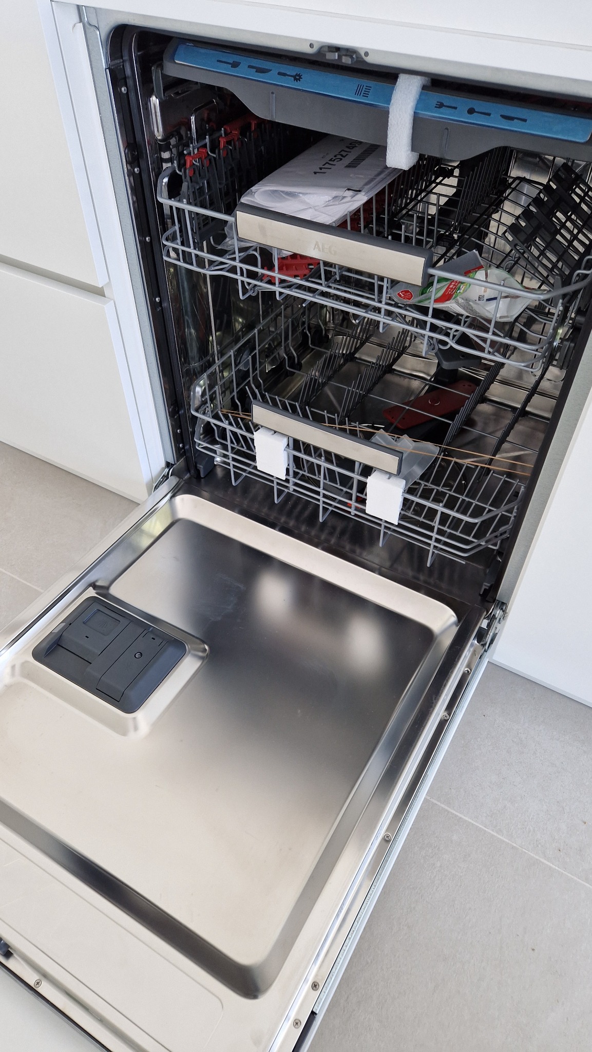 Otwarta, pusta zmywarka marki AEG, w białej zabudowie kuchennej, widoczne metalowe kosze i wnętrze urządzenia.