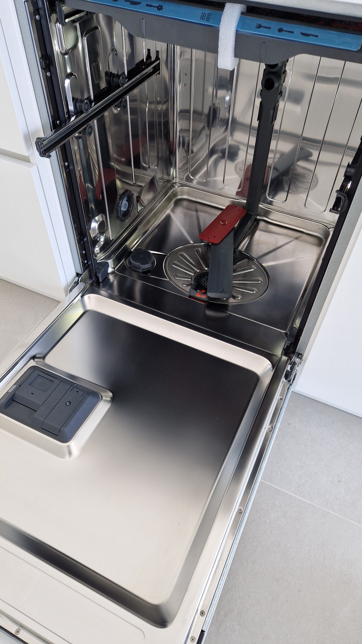 Wnętrze otwartej, czystej zmywarki z widocznymi dyszami spryskującymi i koszem na naczynia, na tle białych szafek kuchennych i jasnej podłogi.