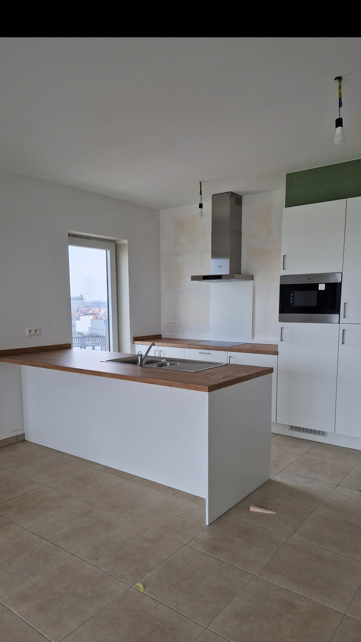 Wnętrze kuchni w stanie surowym: białe szafki, drewniany blat, stalowy okap, ściany przygotowane do malowania, widoczne prowizoryczne oświetlenie i klin pod szafką.