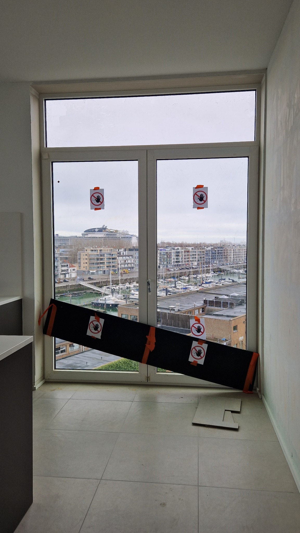 Przygotowanie do mycia okien na wysokości: zabezpieczona szyba z naklejonymi znakami ostrzegawczymi, widok na port jachtowy i zabudowę miejską w tle.