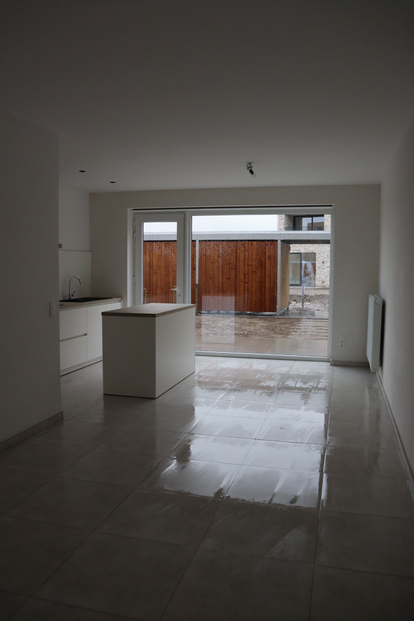 Wnętrze jasnej kuchni z widokiem na mokry plac budowy przez otwarte drzwi balkonowe, świeżo umyta podłoga z połyskiem.