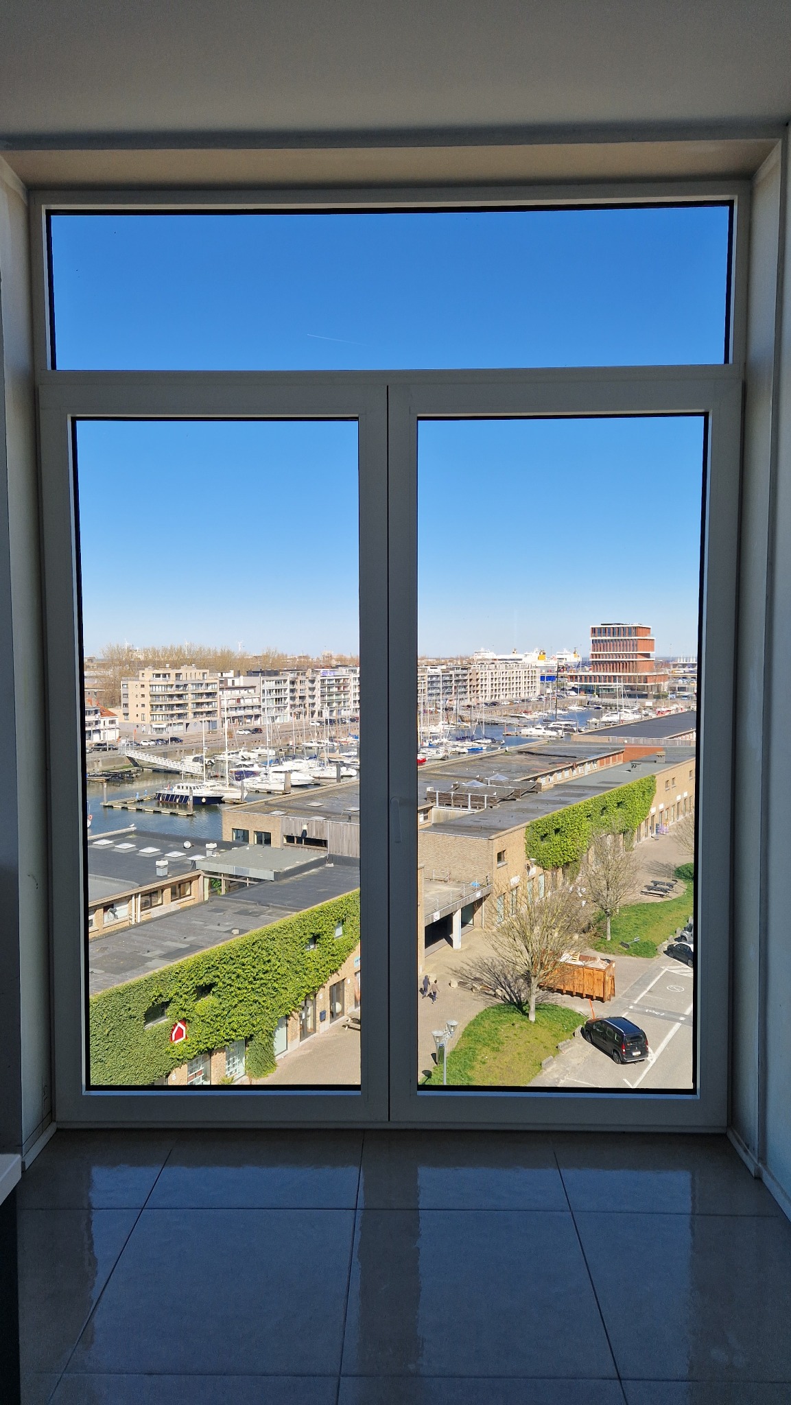 Widok z okna na marinę z jachtami, budynki mieszkalne i biurowe w słoneczny dzień, rama okienna z białego PCV, błyszcząca podłoga z płytek.
