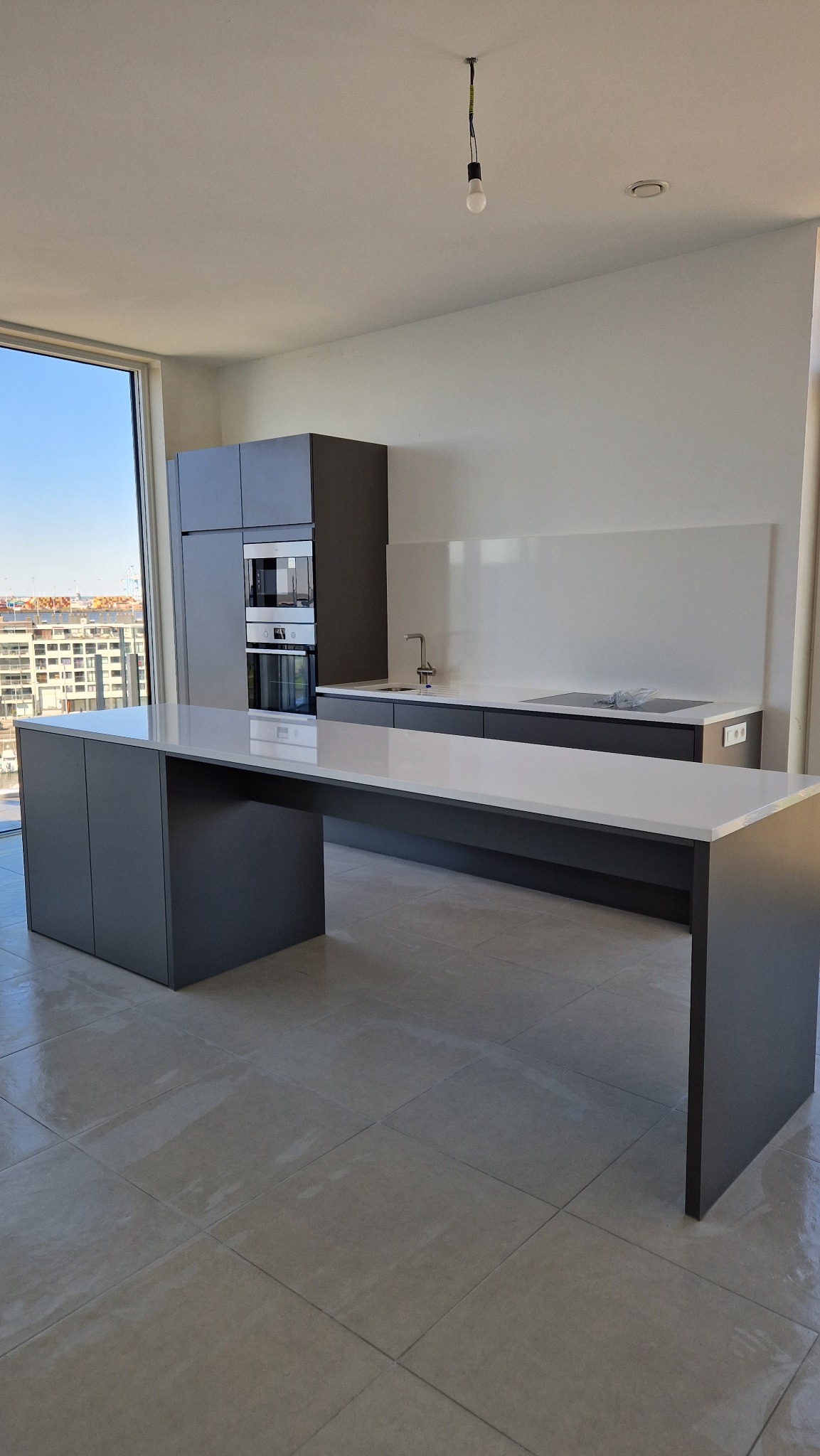 Nowoczesna, minimalistyczna kuchnia w szaro-białej kolorystyce z dużym oknem i widokiem na miasto, ukazująca czyste powierzchnie i uporządkowaną przestrzeń po remoncie.