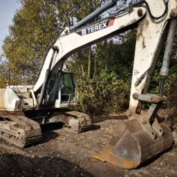 Biała koparka gąsienicowa marki Terex, zaparkowana na ziemi z opadłymi liśćmi, na tle drzew i krzewów w jesiennej scenerii.