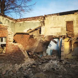 Zrujnowany budynek z czerwonej cegły w trakcie rozbiórki, z widocznymi fragmentami ścian i gruzem, drzewo wyrastające zza ściany, błękitne niebo w tle.