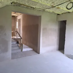 Surowe wnętrze domu w trakcie prac wykończeniowych: ściany pokryte świeżym tynkiem, widoczne schody z drewnianą barierką, izolacja piankowa na suficie, pojedynczy bloczek betonowy na podłodze.