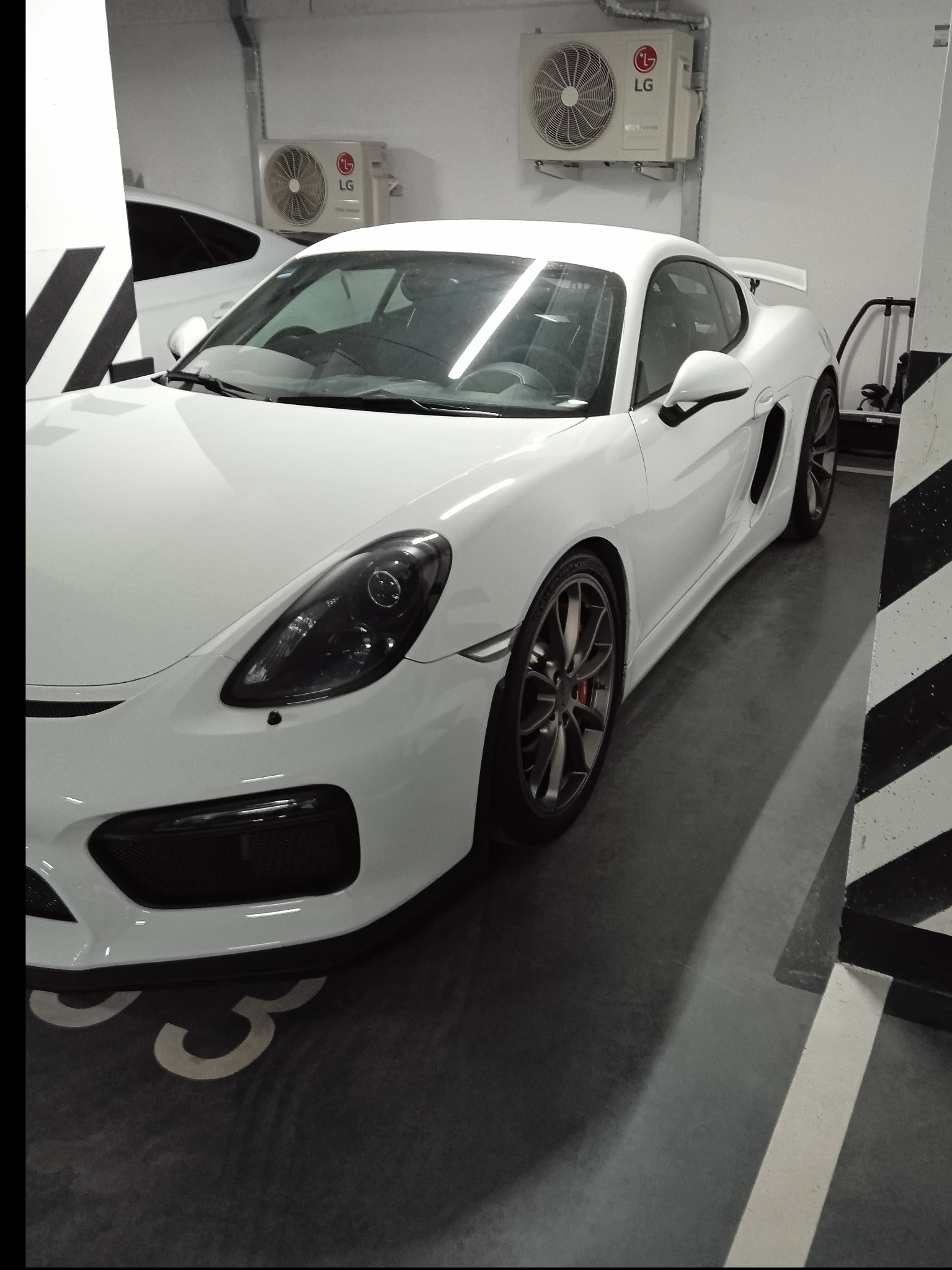 Biały sportowy samochód Porsche GT4 zaparkowany w garażu podziemnym, widoczne czarne pasy na filarze i klimatyzatory LG na ścianie w tle.