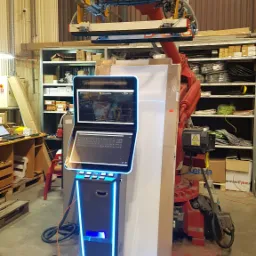 Roboczy widok stanowiska z robotem przemysłowym, panelem sterowania z ekranem i klawiaturą, oraz elementami oświetlenia LED w hali produkcyjnej.