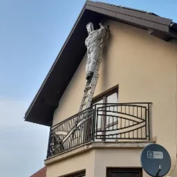 Pracownik w kombinezonie ochronnym na drabinie, wykonujący prace na wysokości przy elewacji budynku z balkonem.