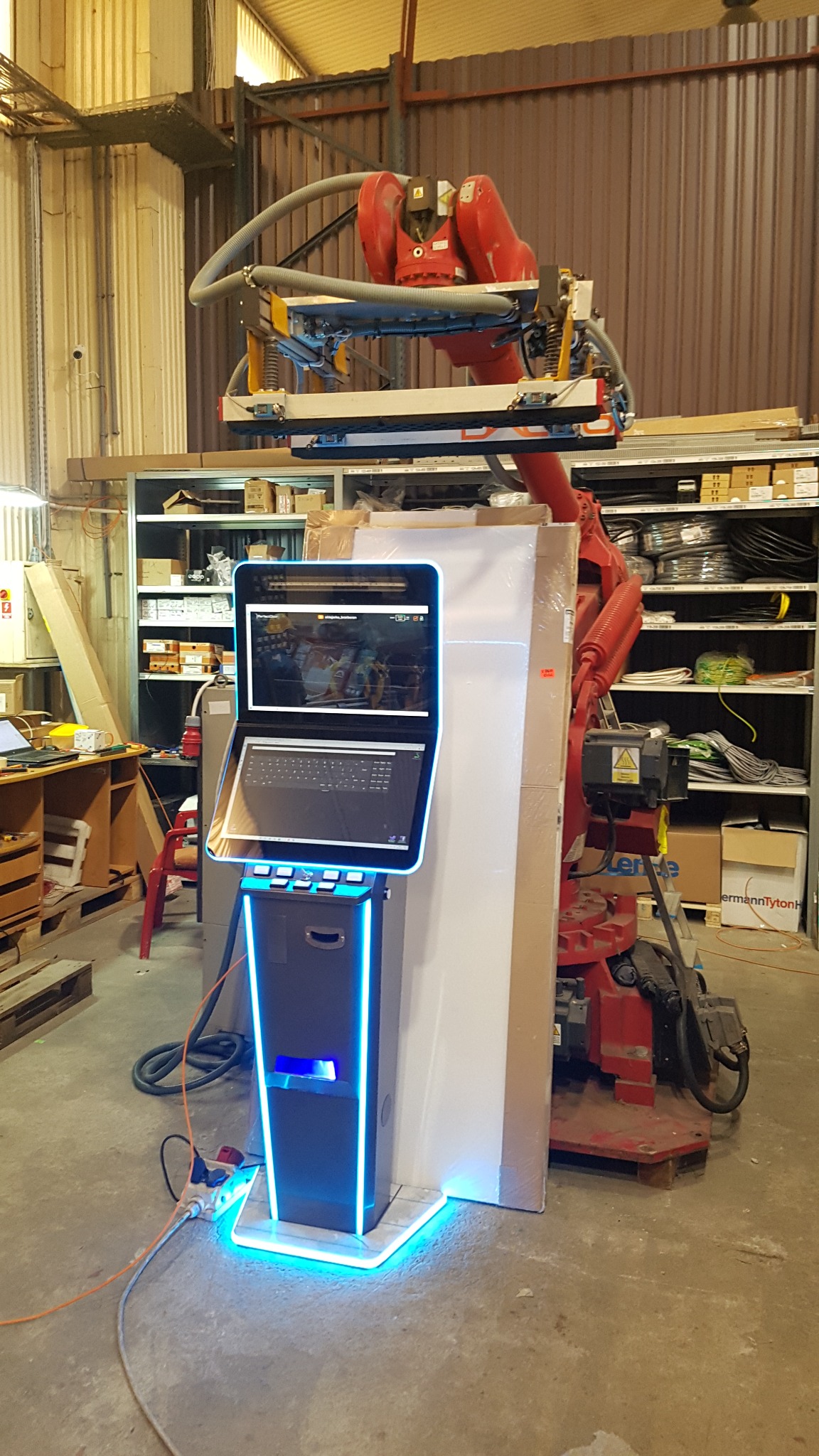 Roboczy widok stanowiska z robotem przemysłowym, panelem sterowania z ekranem i klawiaturą, oraz elementami oświetlenia LED w hali produkcyjnej.