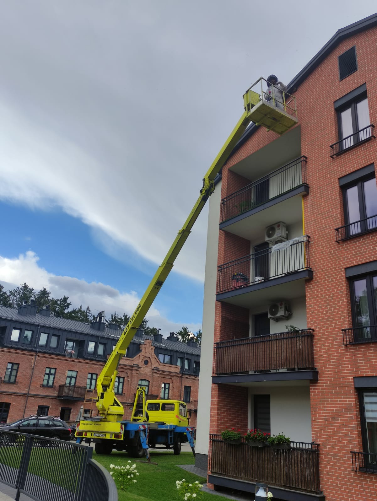 Pracownik w kombinezonie ochronnym na podnośniku koszowym, usuwa gniazdo owadów z elewacji budynku mieszkalnego z cegły.