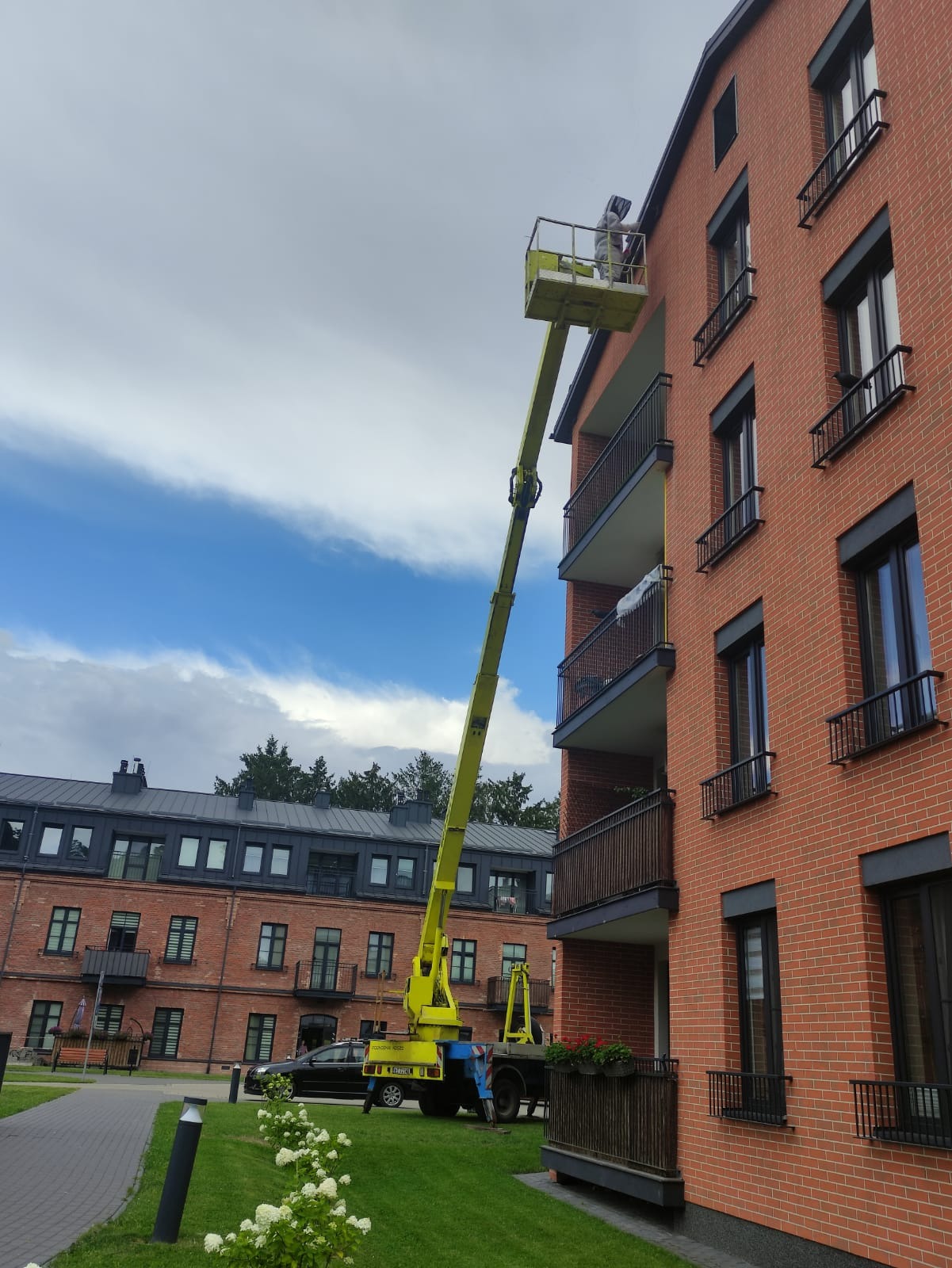 Pracownik w podnośniku koszowym wykonuje prace na elewacji budynku z czerwonej cegły, widoczne balkony z ciemną balustradą, w tle budynek z szarym dachem i zielenią.