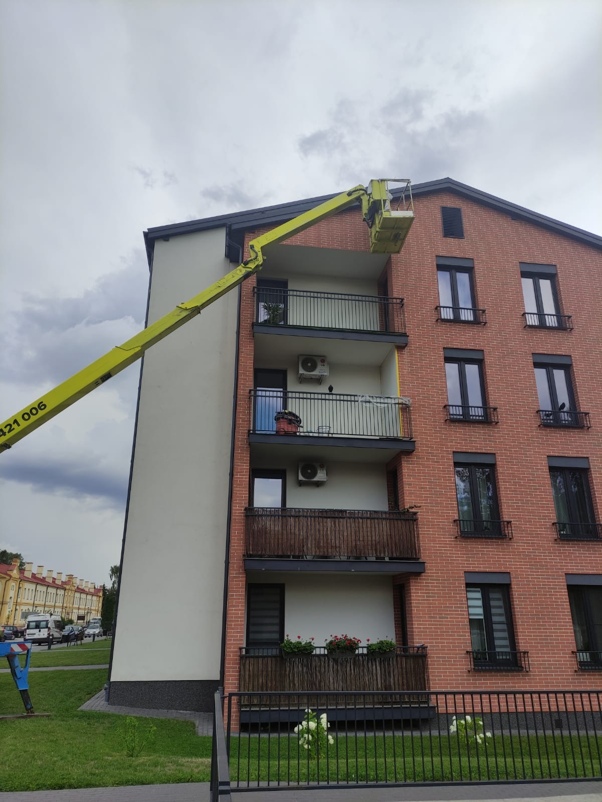 Żółty podnośnik koszowy przy elewacji budynku mieszkalnego z cegły, balkony z klimatyzatorami, widok z dołu na pochmurne niebo.