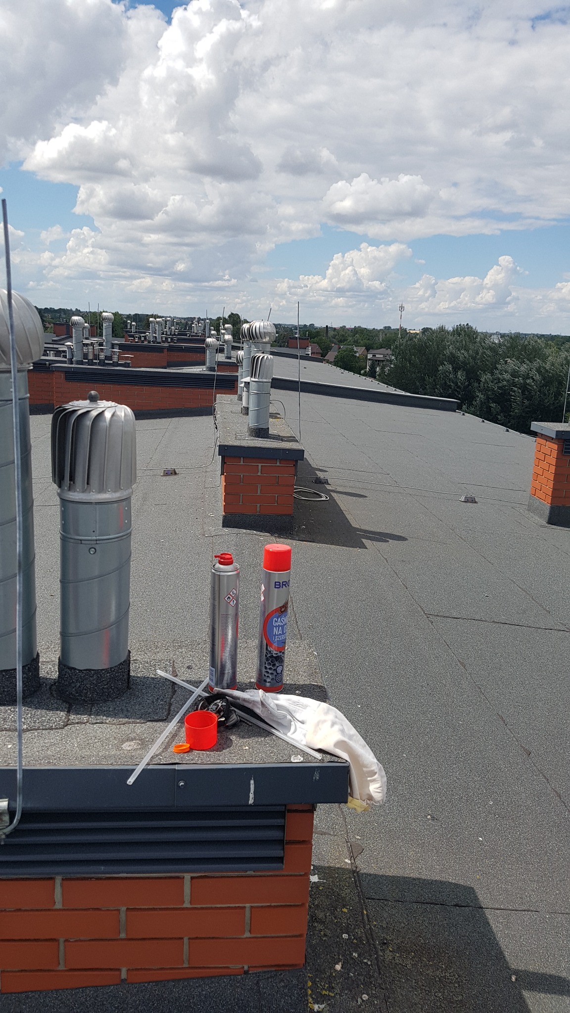 Widok z góry na płaski dach z licznymi kominami wentylacyjnymi, na pierwszym planie dwie puszki aerozolu, rękawice robocze i drobne narzędzia, w tle błękitne niebo z chmurami i zabudowa miejska.