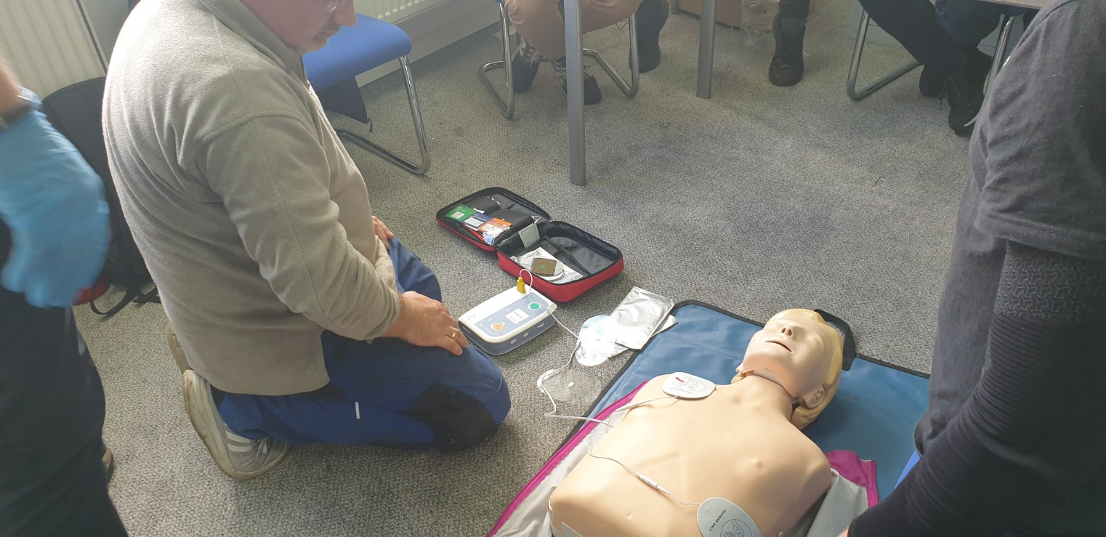 Instruktor demonstruje użycie defibrylatora na fantomie podczas zajęć z pierwszej pomocy, widoczna otwarta torba z akcesoriami i elektrody.