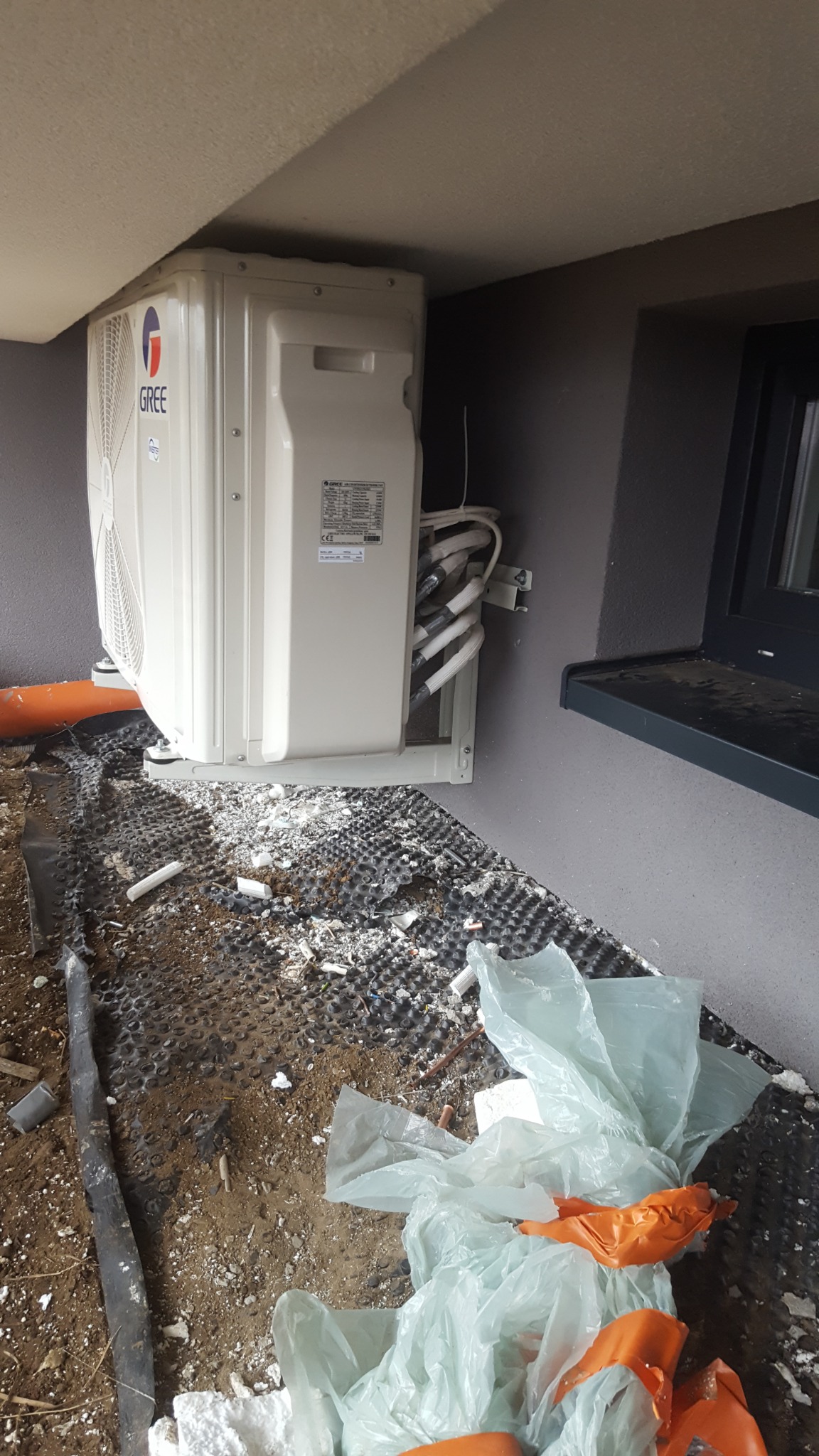 Biała jednostka zewnętrzna klimatyzatora firmy Gree zamontowana na ścianie budynku, widoczne rury i kable, na podłodze gruz i folie budowlane.