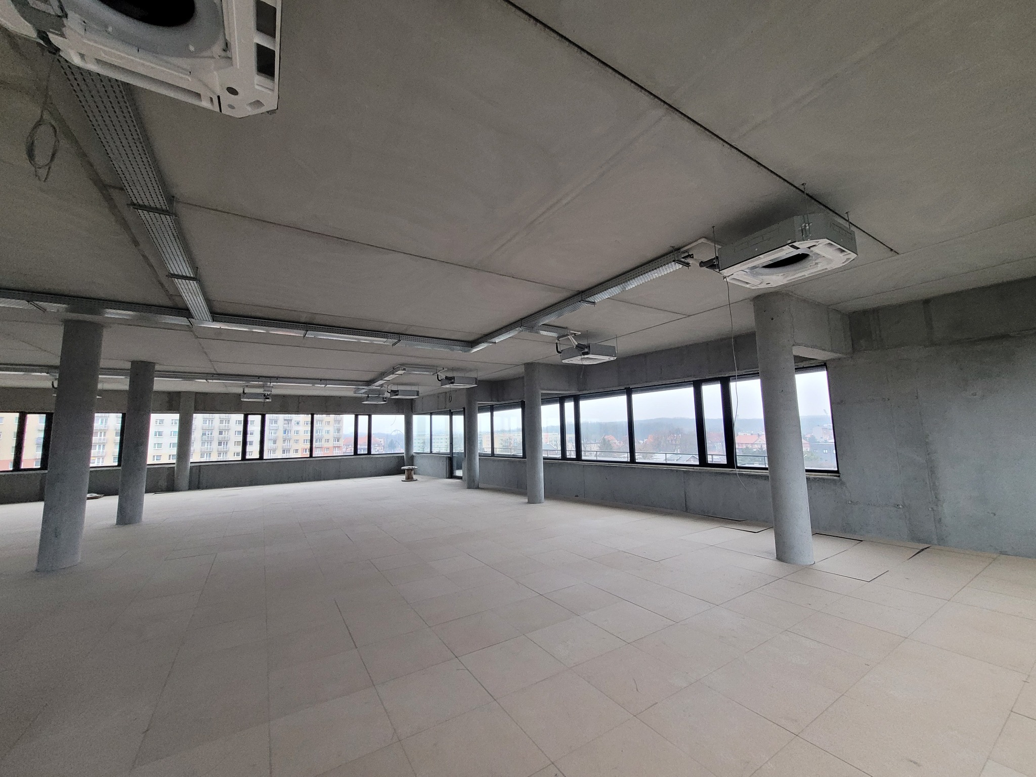 Wnętrze biurowe w stanie deweloperskim z widocznymi elementami wentylacji pod sufitem, betonowe filary i surowy wystrój, duże okna z widokiem na miasto.