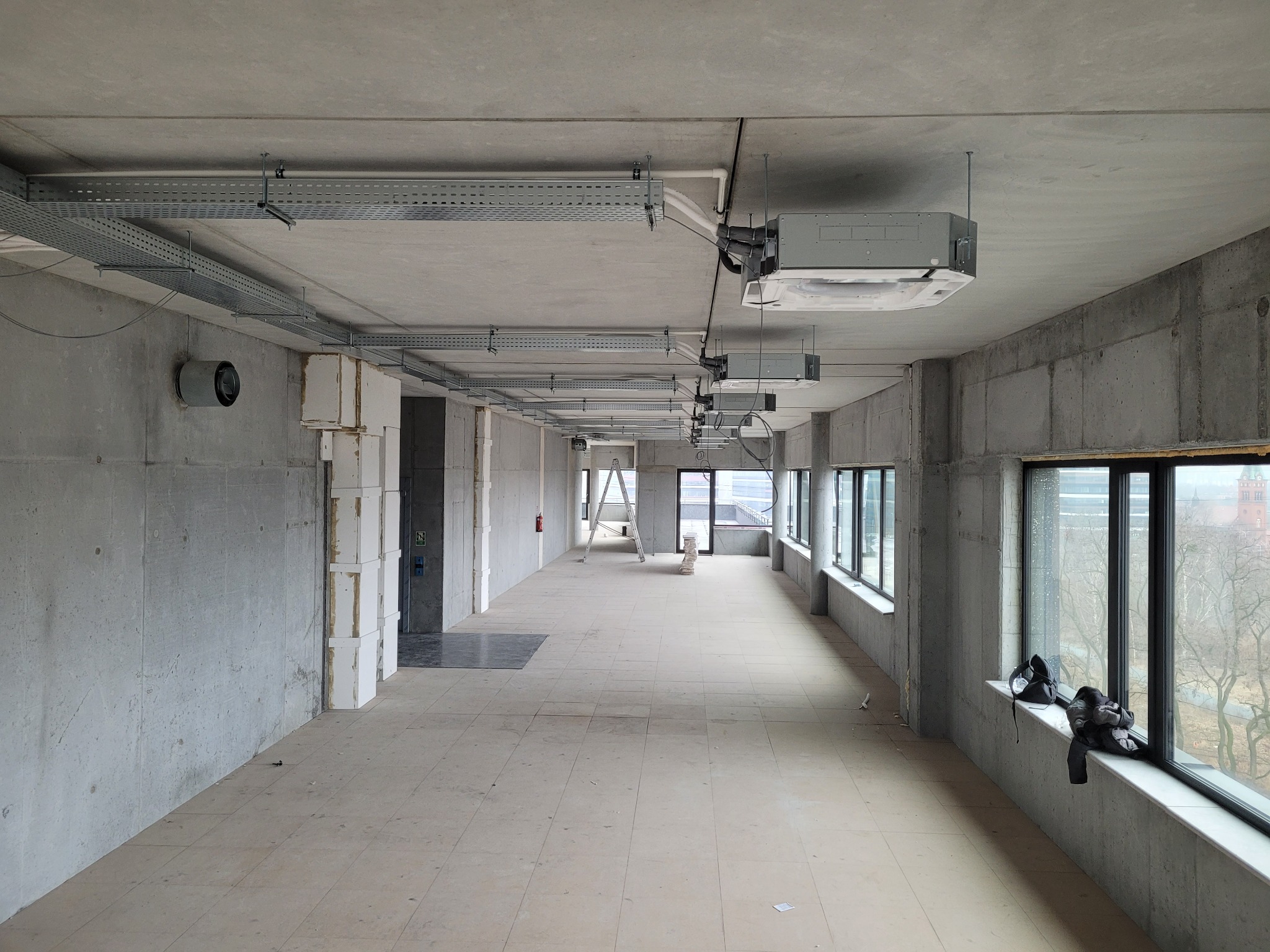 Wnętrze surowego biura w trakcie wykańczania: widoczne kanały wentylacyjne pod sufitem, klimatyzatory kasetonowe, betonowe ściany i podłoga z płytek, drabina w oddali oraz ubrania na parapecie okna.