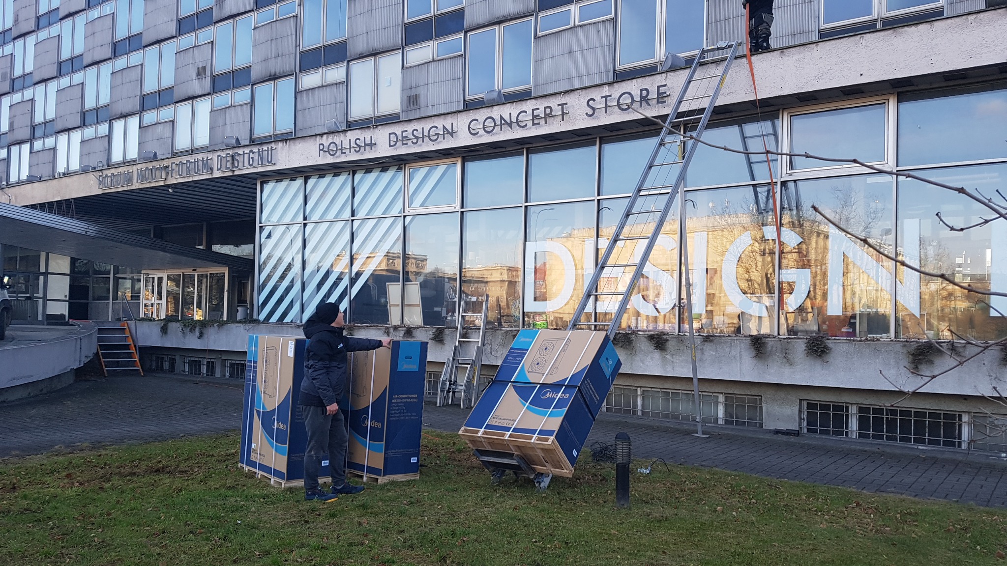 Dostawa fabrycznie nowych klimatyzatorów Midea do budynku Polish Design Concept Store; mężczyzna w czapce wskazuje na jedno z pudeł, a w tle drabina oparta o elewację budynku.