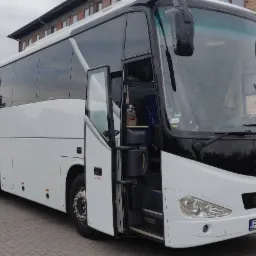 Biały autokar z otwartymi drzwiami na parkingu, widoczny numer rejestracyjny i budynek z cegły w tle.