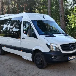 Biały bus Mercedes-Benz Sprinter z przyciemnianymi szybami, zaparkowany na leśnym parkingu obok znaku drogowego z ograniczeniem prędkości do 30 km/h i symbolem parkingu dla osób niepełnosprawnych.