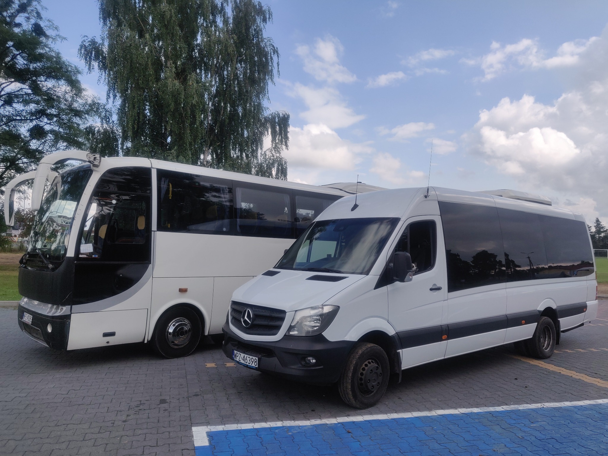 Dwa białe busy, Mercedes Sprinter i autokar, zaparkowane na parkingu z kostki brukowej z niebieskimi liniami, drzewa w tle, pochmurne niebo.
